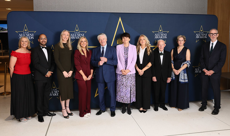 A group of ten award winners and presenters stand together in front of the UCD Alumni Awards 2025 backdrop, all dressed in formal attire.